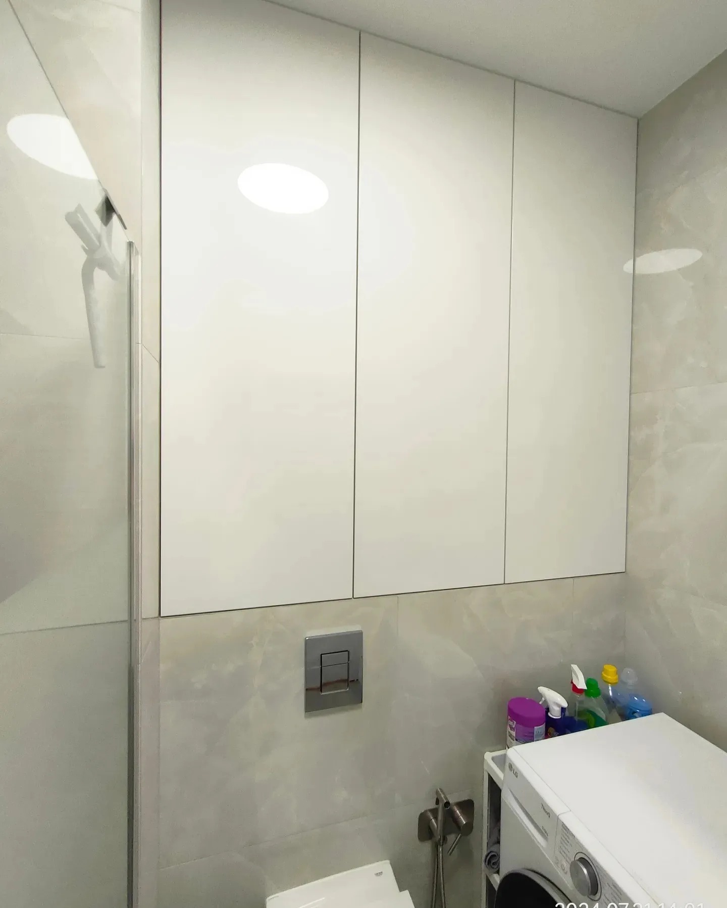 Białe szafki wiszące w łazience nad pralką i toaletą, z widocznymi środkami czystości i armaturą.