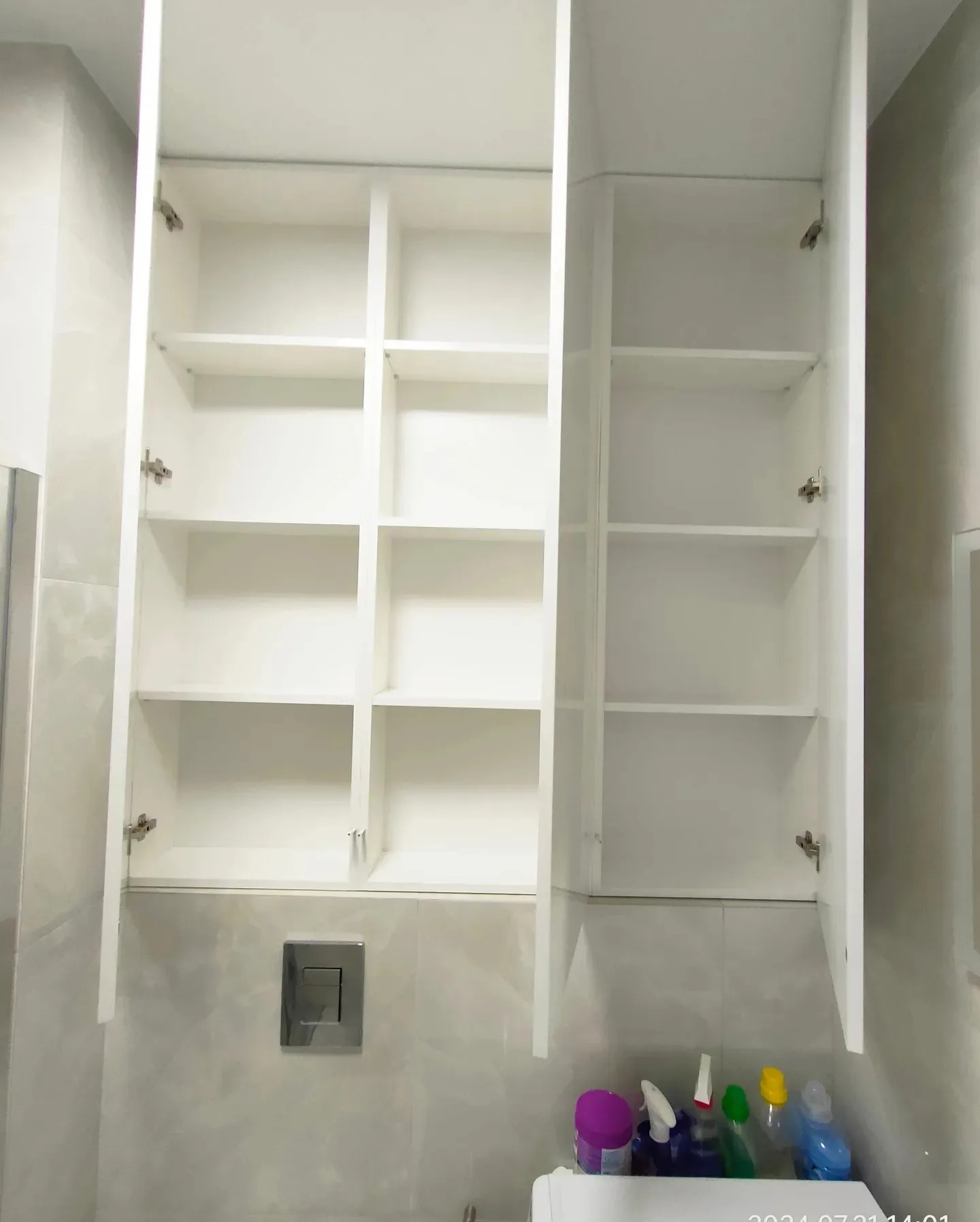 Biała, otwarta szafka łazienkowa z półkami, zamontowana nad toaletą podwieszaną z widocznym przyciskiem spłukiwania. Na dole widoczne środki czystości.