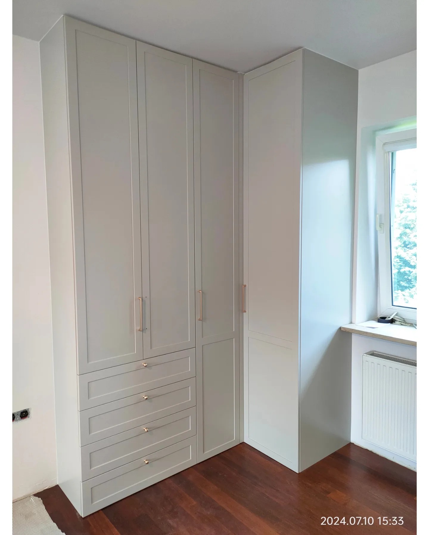 Jasnoszara szafa narożna z czterema szufladami i długimi, smukłymi uchwytami, dopasowana do rogu pokoju z drewnianą podłogą i oknem.