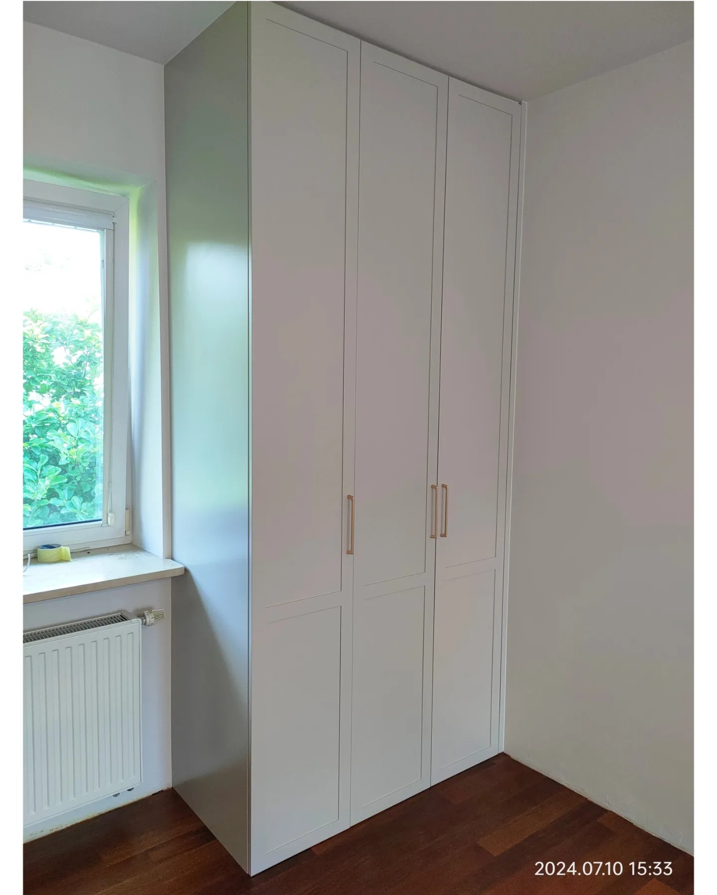 Biała, wysoka szafa z trzema segmentami, zintegrowana z wnęką w pokoju, z widocznym oknem i grzejnikiem.