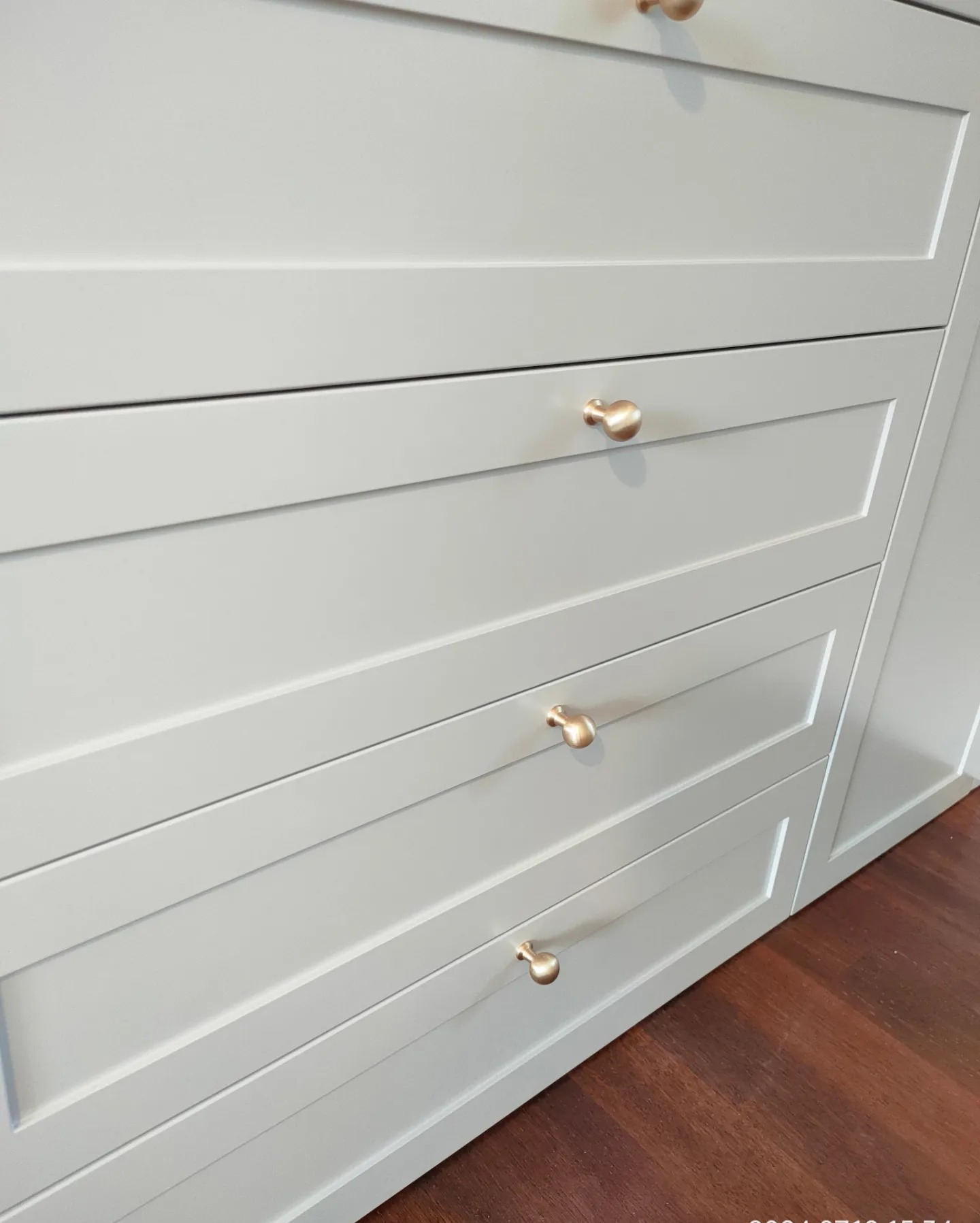 Jasnoszare szuflady z frezowanymi frontami i złotymi gałkami na tle ciemnej, drewnianej podłogi.