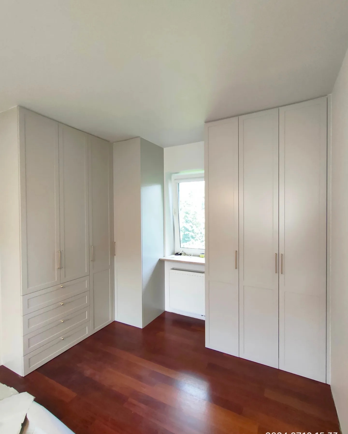 Jasnoszare szafy wnękowe z szufladami i złotymi uchwytami, dopasowane do wnęki w pokoju z drewnianą podłogą i oknem.