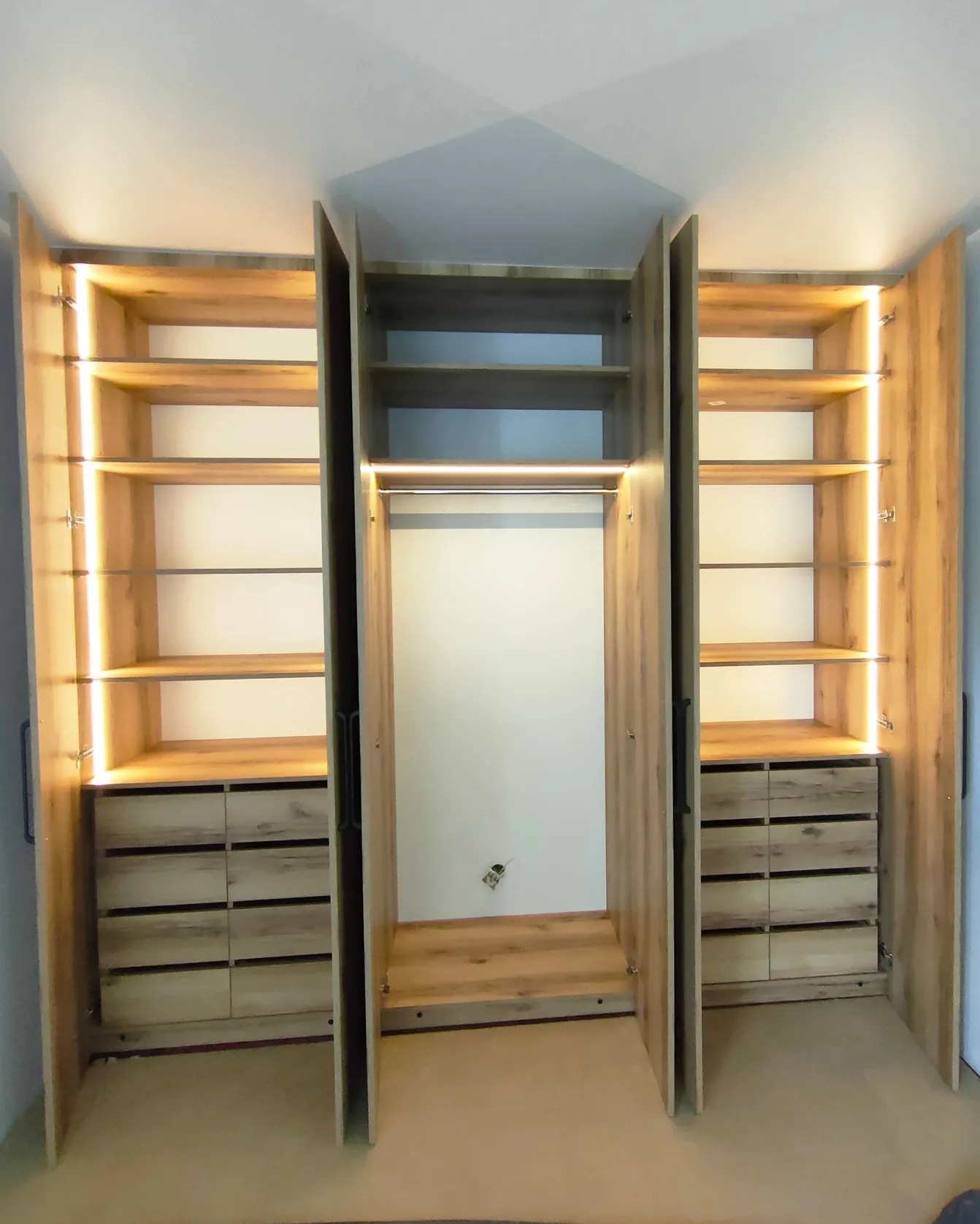 Otwarta szafa z oświetleniem LED wewnątrz, widoczne półki i szuflady z drewnianym wzorem, trzy segmenty z drzwiami uchylnymi.