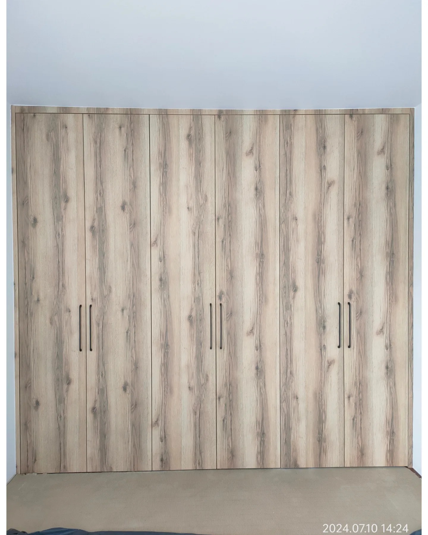 Duża szafa wnękowa z ośmioma drzwiami o jasnym, drewnopodobnym wykończeniu z pionowymi słojami i czarnymi, minimalistycznymi uchwytami.