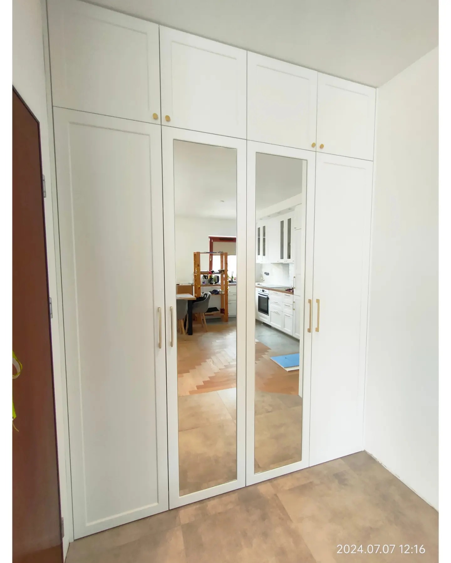 Biała szafa wnękowa z lustrzanymi drzwiami, odbijająca wnętrze mieszkania z widoczną kuchnią i jadalnią.