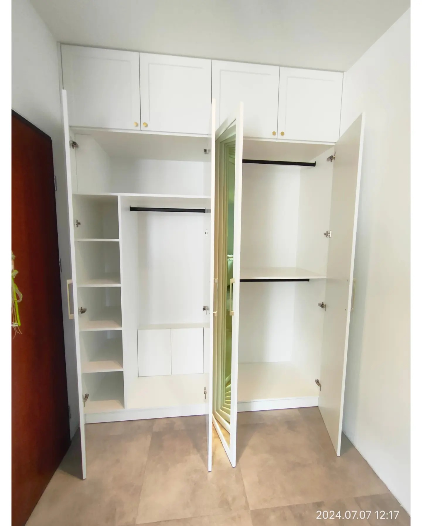 Biała szafa wnękowa z otwartymi drzwiami, widoczne półki i drążki, górne szafki z małymi złotymi uchwytami, na podłodze płytki w odcieniach beżu.