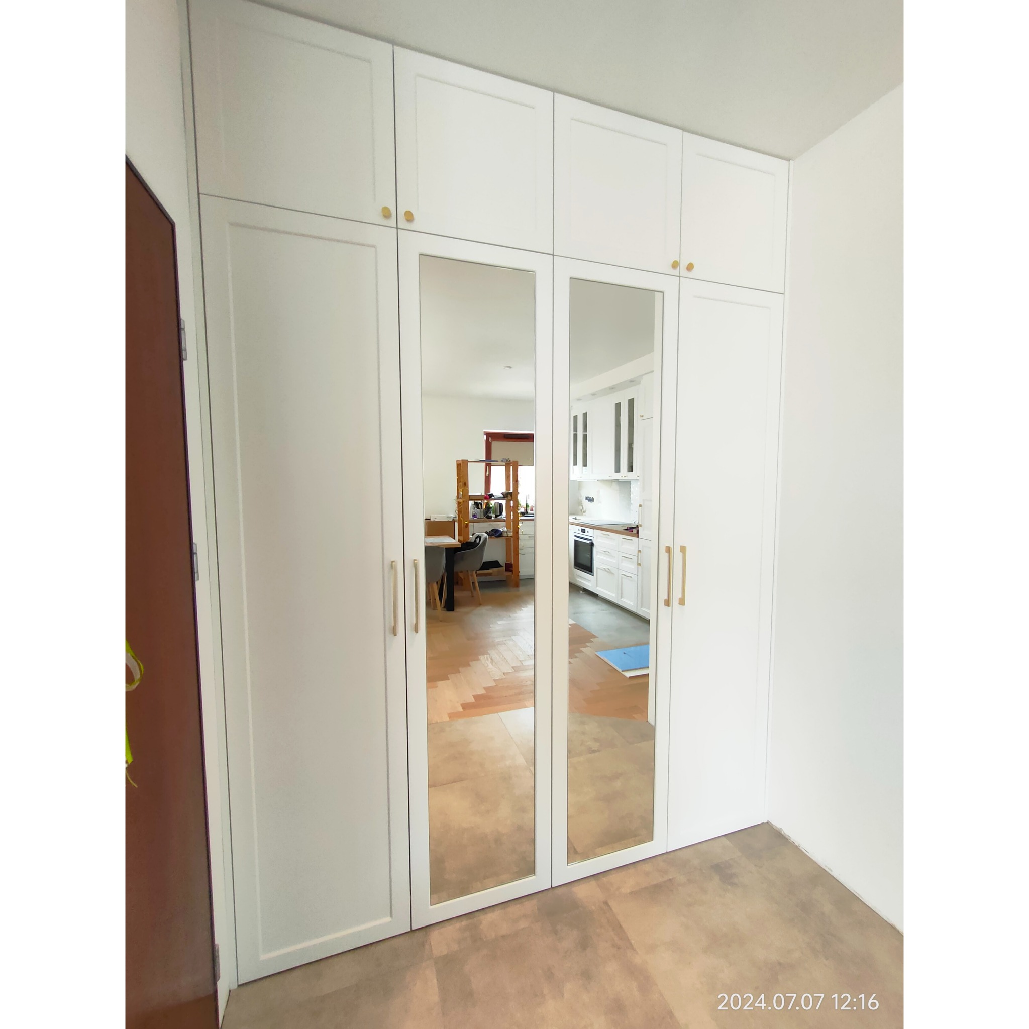 Biała szafa wnękowa z lustrzanymi drzwiami, widoczne złote uchwyty i odbicie kuchni w lustrze.