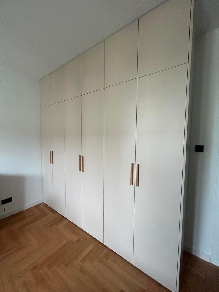 Biała zabudowa meblowa od podłogi do sufitu z drewnianymi uchwytami, parkiet w jodełkę na podłodze.