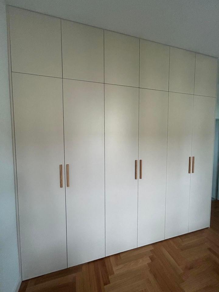 Pełna ściana zabudowana szafą z jasnymi frontami i minimalistycznymi, drewnianymi uchwytami, na podłodze parkiet ułożony w jodełkę.