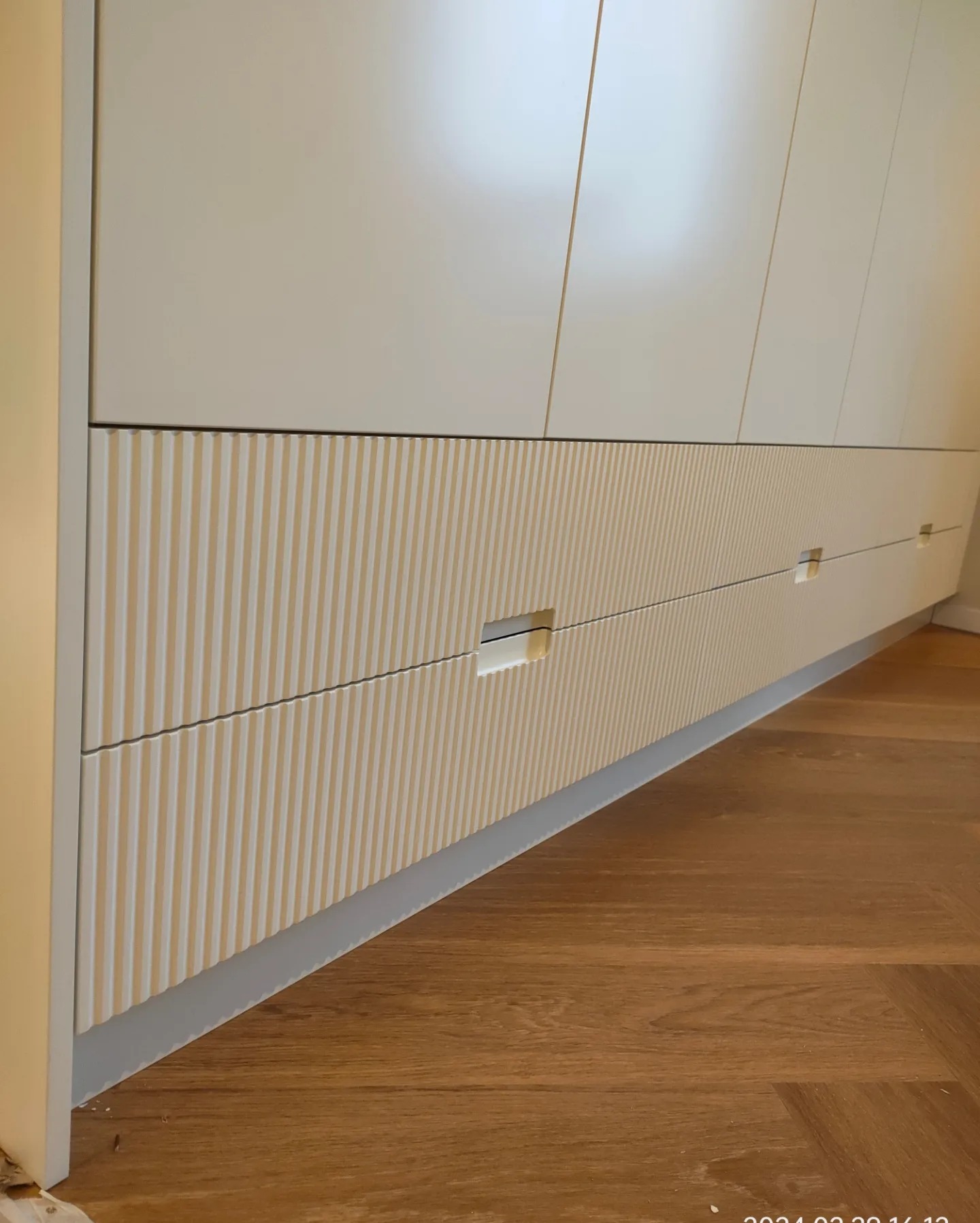 Biała szafa z ryflowanymi frontami szuflad i ukrytymi uchwytami, widok z dołu na drewnianą podłogę.