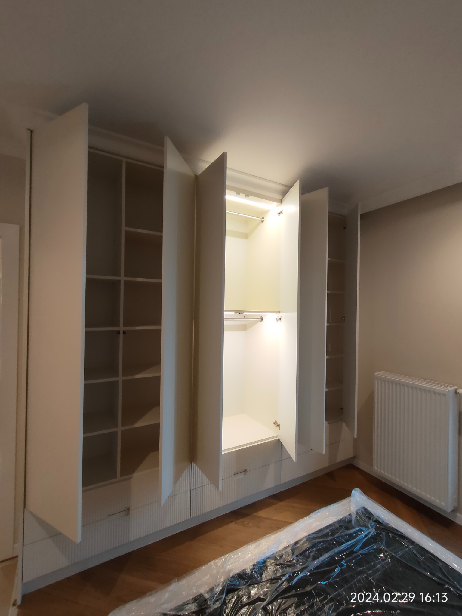 Wnętrze szafy wnękowej z otwartymi drzwiami, podświetlane półki i drążek na ubrania, widoczne elementy wykończenia z ryflowanymi frontami, na podłodze folia ochronna.