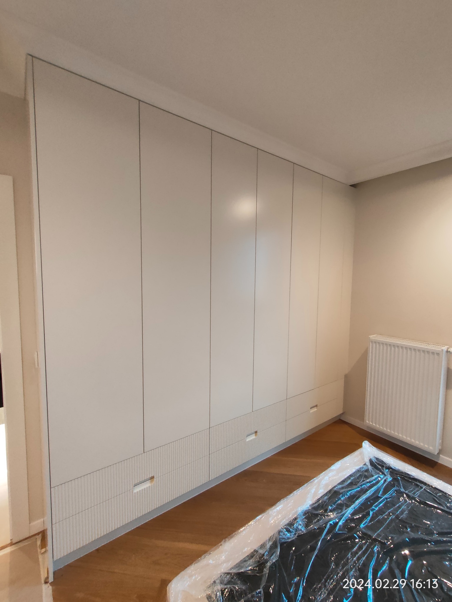 Wysoka, biała szafa wnękowa z frezowanymi frontami szuflad w dolnej części, zrealizowana w mieszkaniu w Warszawie. Na podłodze folia ochronna.