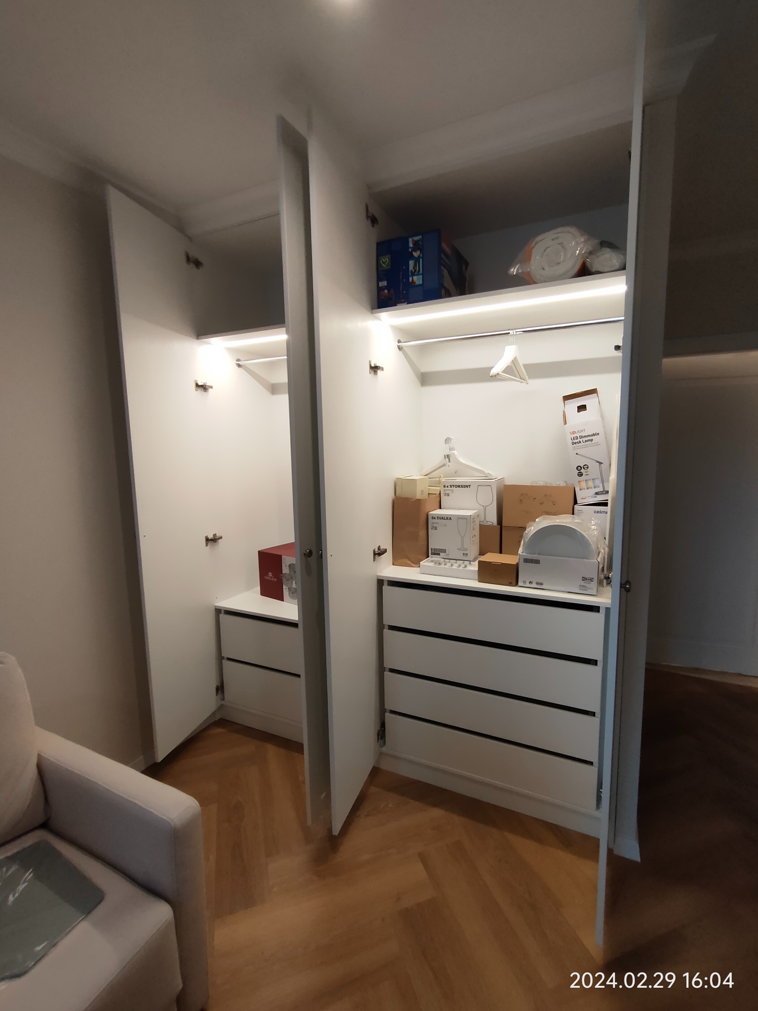 Białe szafy z szufladami, częściowo otwarte, z oświetleniem LED i zawartością w postaci pudełek i wieszaków, na podłodze parkiet ułożony w jodełkę.