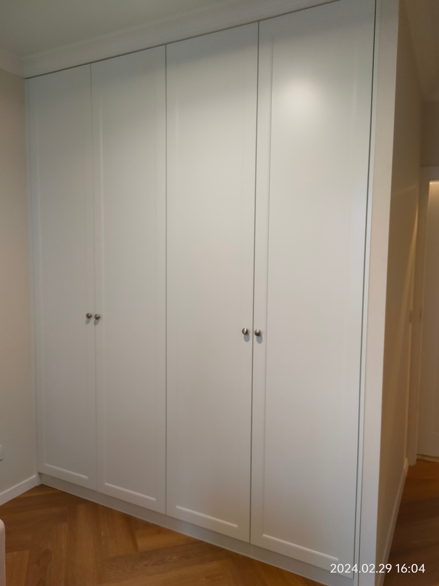 Biała, czterodrzwiowa szafa wnękowa z minimalistycznymi, okrągłymi uchwytami, dopasowana do listwy przypodłogowej i sufitu, na podłodze parkiet w jodełkę.