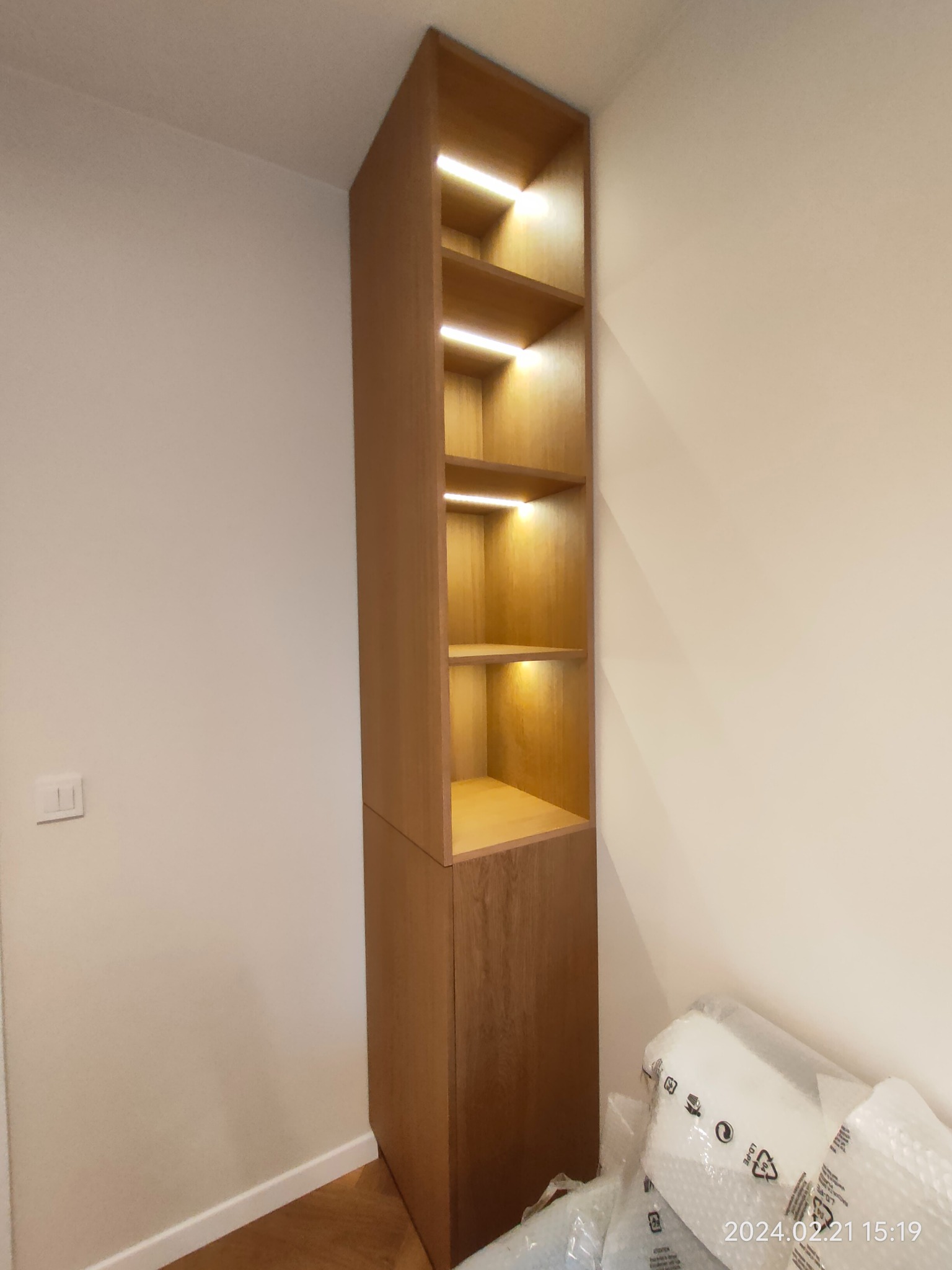 Wysoka, drewniana szafka wnękowa z oświetleniem LED na półkach, umieszczona w rogu pokoju, z widocznymi elementami opakowania po montażu.