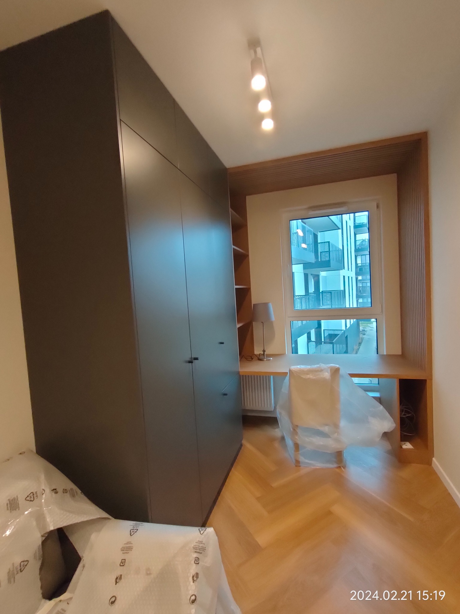 Ciemnoszara szafa wnękowa z biurkiem i regałem wbudowanym w ścianę, krzesło zabezpieczone folią ochronną, parkiet w jodełkę.