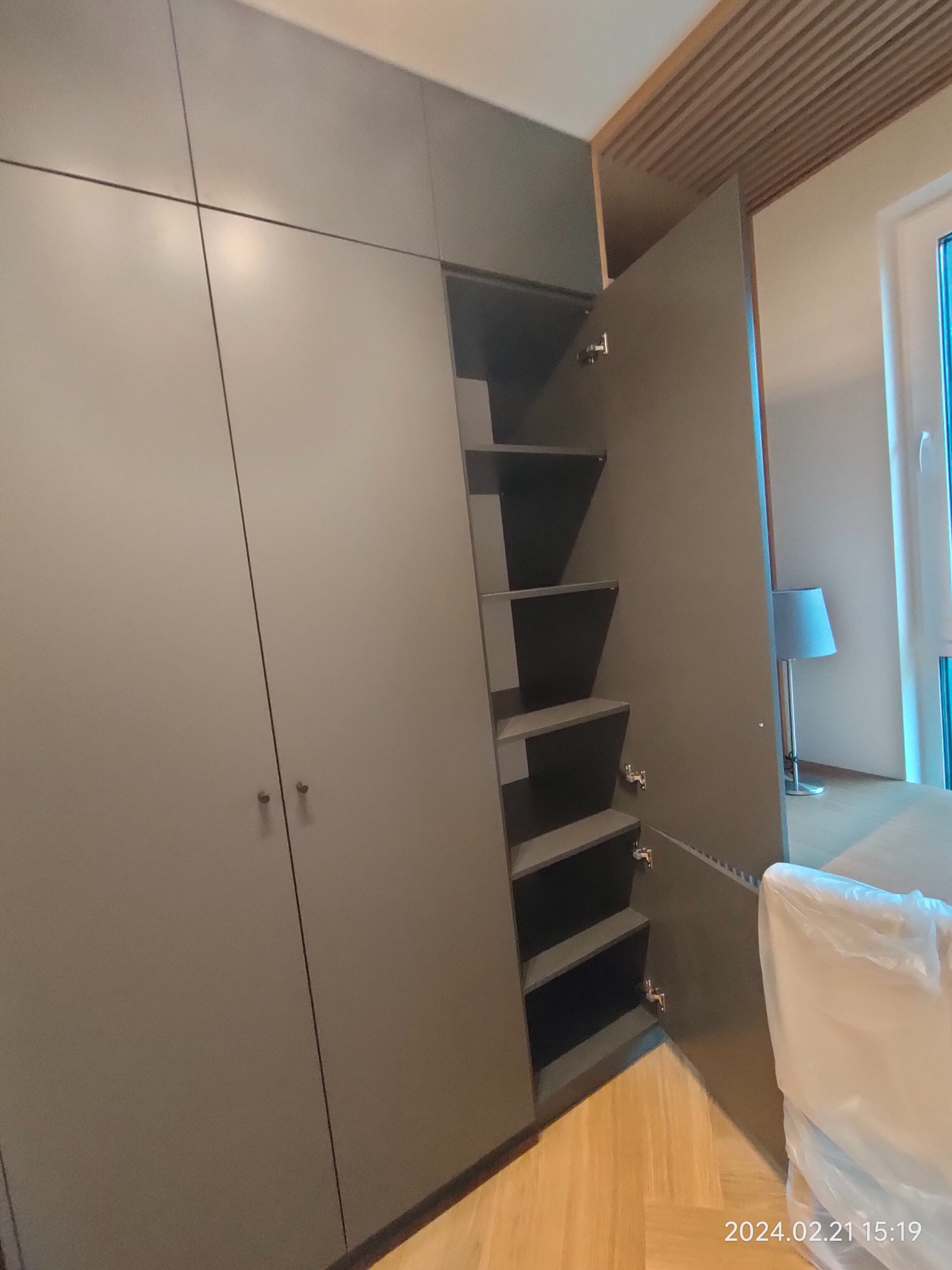 Szara szafa z otwartymi drzwiami ukazującymi półki, minimalistyczny design, drewniany sufit w tle, parkiet na podłodze.