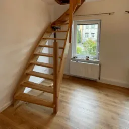 Drewniane schody z naturalnego drewna, z widocznymi sękami, prowadzące na poddasze, zamontowane w jasnym pomieszczeniu z panelami podłogowymi i dwoma oknami, na parapecie jednego z nich widoczna...