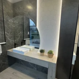 Nowoczesna łazienka z marmurowymi płytkami, umywalkami nablatowymi, lustrem z podświetleniem LED i dekoracyjnymi roślinami w doniczkach.