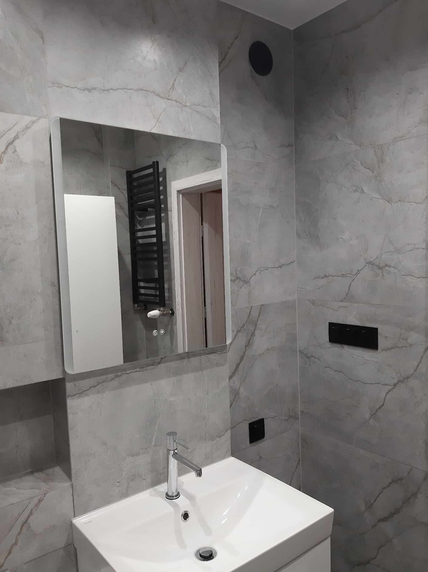 Nowoczesna łazienka z szarymi płytkami imitującymi marmur, białą umywalką nablatową i chromowaną baterią. W lustrze odbija się grzejnik drabinkowy i biała szafka. Widoczny fragment drzwi do innego...