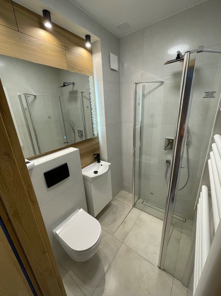 Nowoczesna łazienka z kabiną prysznicową, wiszącą toaletą i małą umywalką, wykończona w jasnych szarościach i bieli, z drewnianymi akcentami nad lustrem.