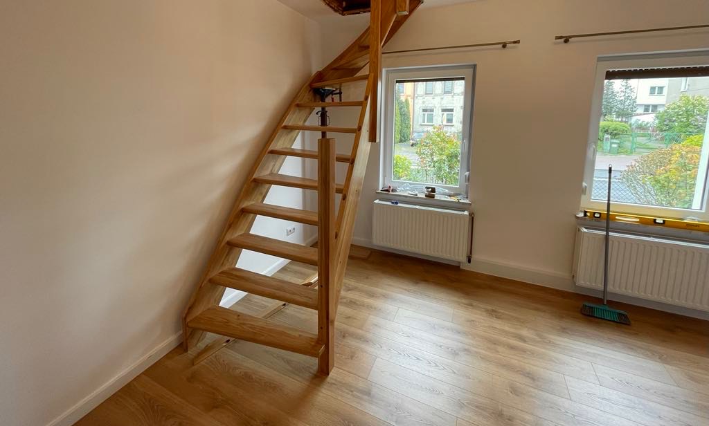 Drewniane schody z naturalnego drewna, z widocznymi sękami, prowadzące na poddasze, zamontowane w jasnym pomieszczeniu z panelami podłogowymi i dwoma oknami, na parapecie jednego z nich widoczna...