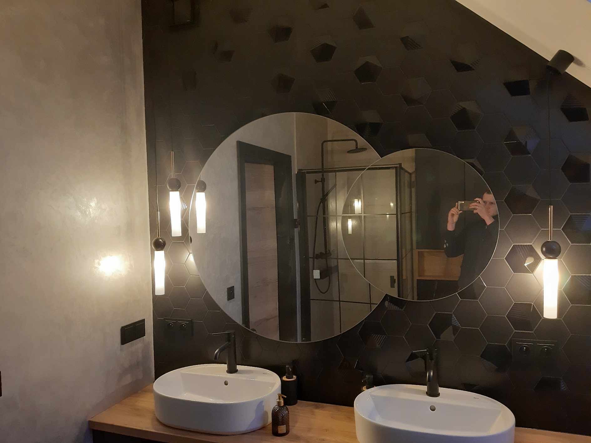 Nowoczesna łazienka z dwoma umywalkami nablatowymi, czarnymi heksagonalnymi płytkami na ścianie i okrągłymi lustrami, w których odbija się osoba robiąca zdjęcie