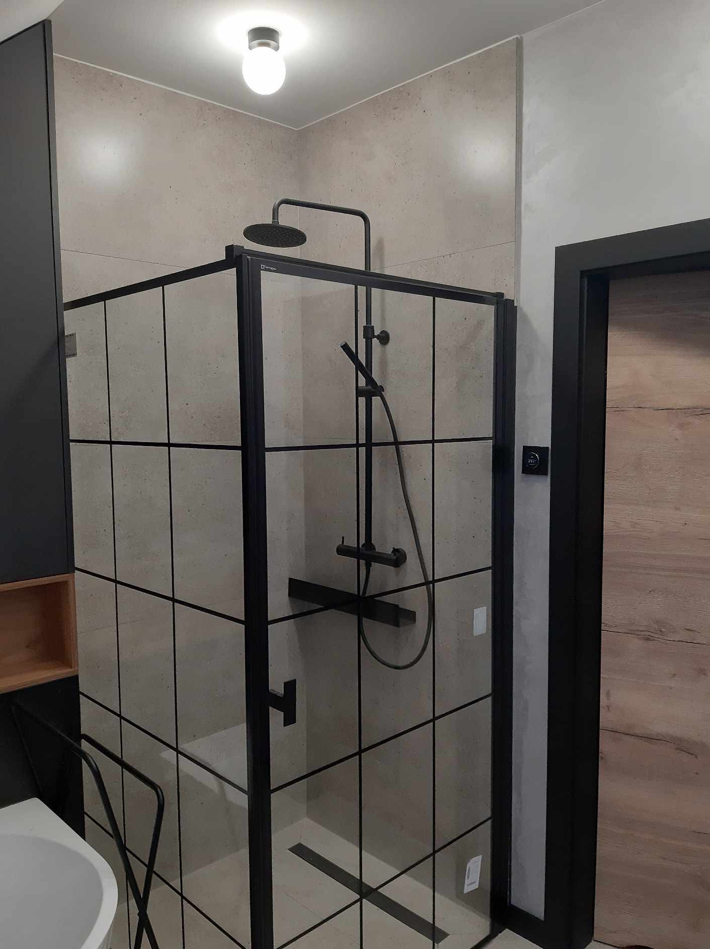 Nowoczesna kabina prysznicowa z czarnymi profilami i deszczownicą, szare płytki imitujące beton, minimalistyczne oświetlenie sufitowe.