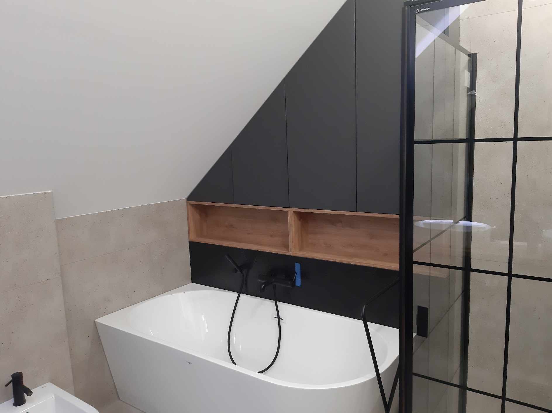 Nowoczesna łazienka na poddaszu z białą wanną, czarną armaturą i zabudową szafek pod skosem w kolorze grafitowym z drewnianymi wnękami, kabina prysznicowa z czarnymi szprosami.