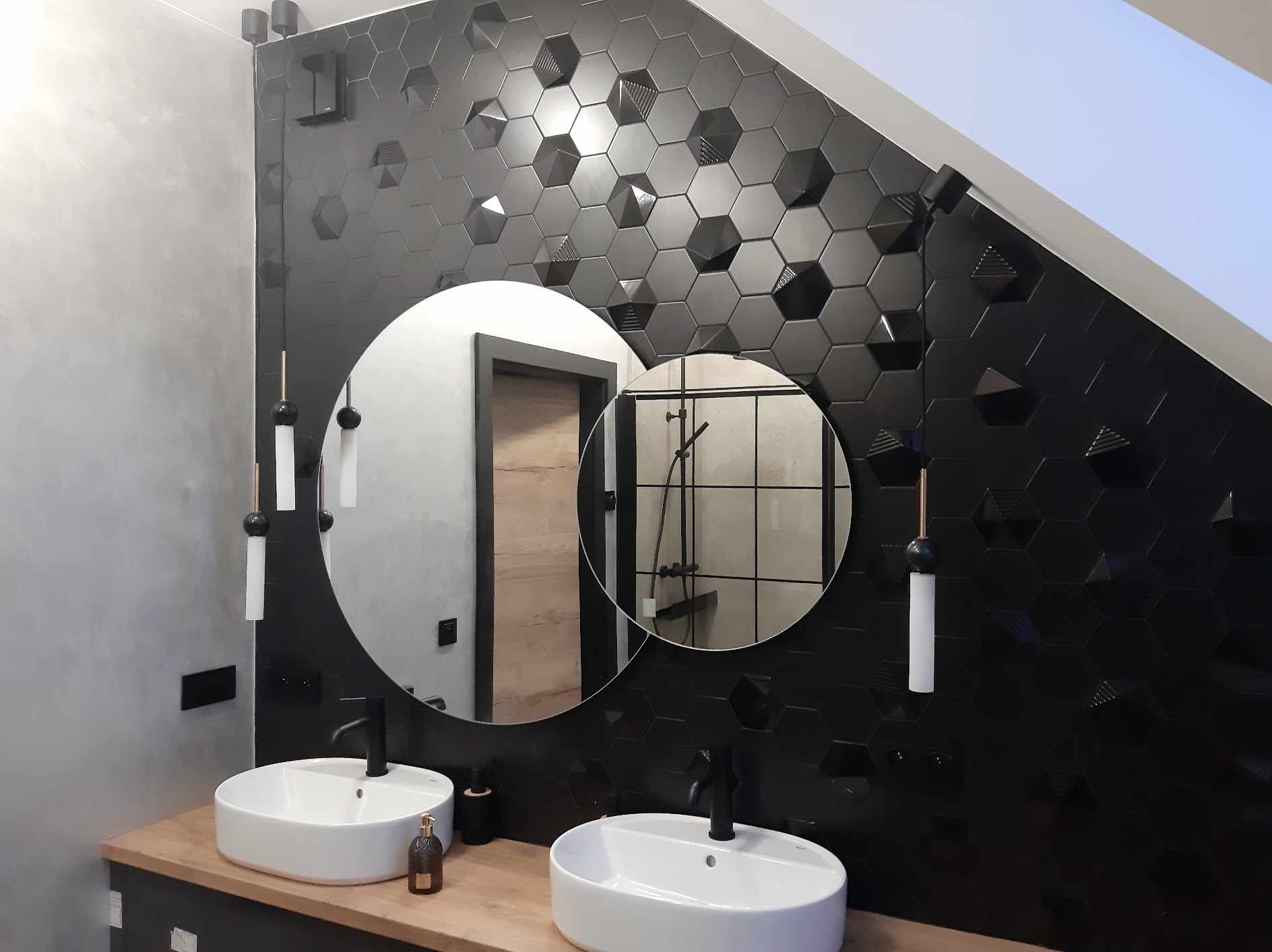 Nowoczesna łazienka z czarnymi, heksagonalnymi płytkami 3D na ścianie, okrągłymi lustrami i białymi umywalkami nablatowymi na drewnianym blacie.