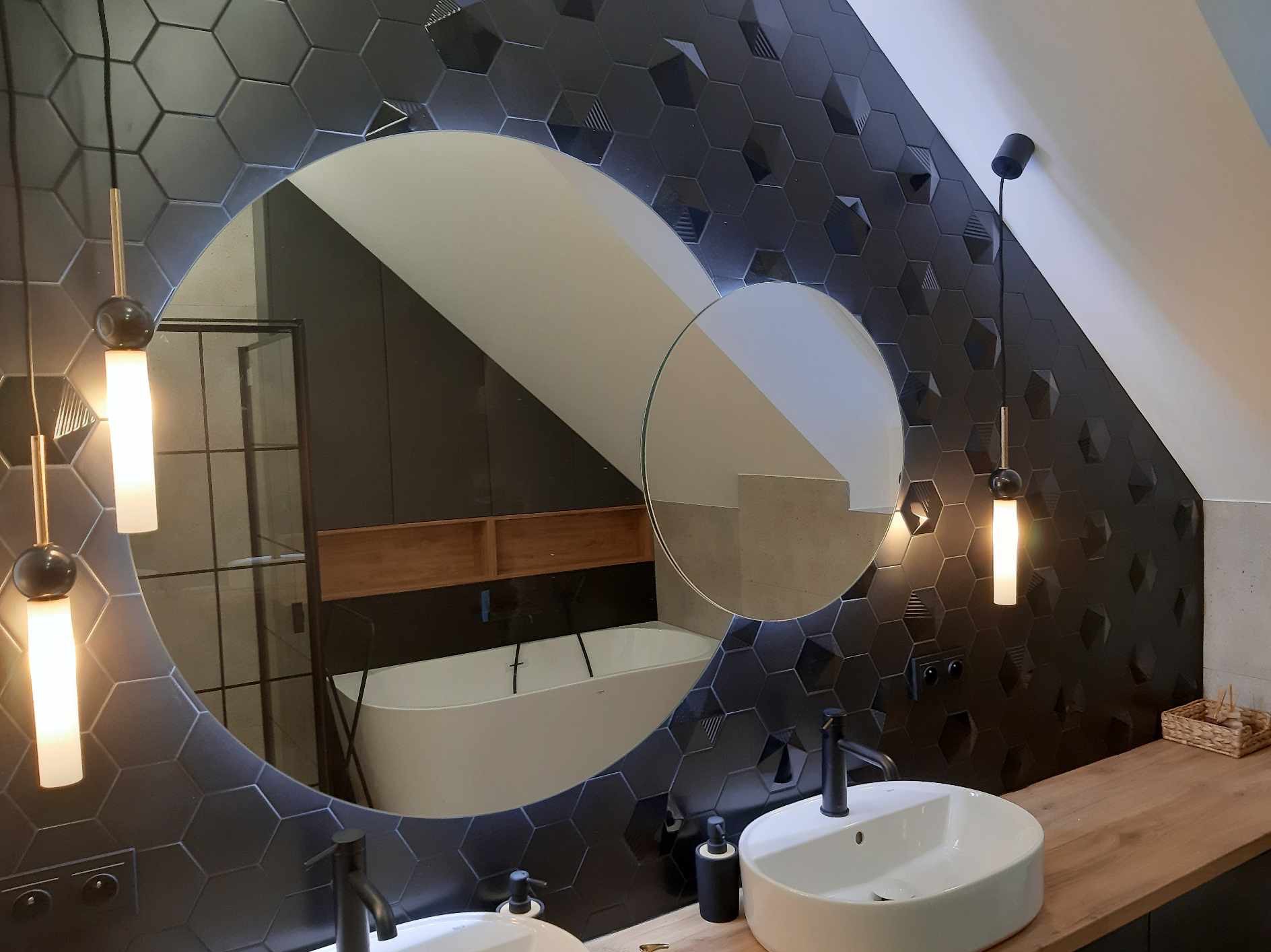 Nowoczesna łazienka na poddaszu z czarnymi heksagonalnymi płytkami 3D, okrągłym lustrem odbijającym wannę, drewnianym blatem i wiszącymi lampami.