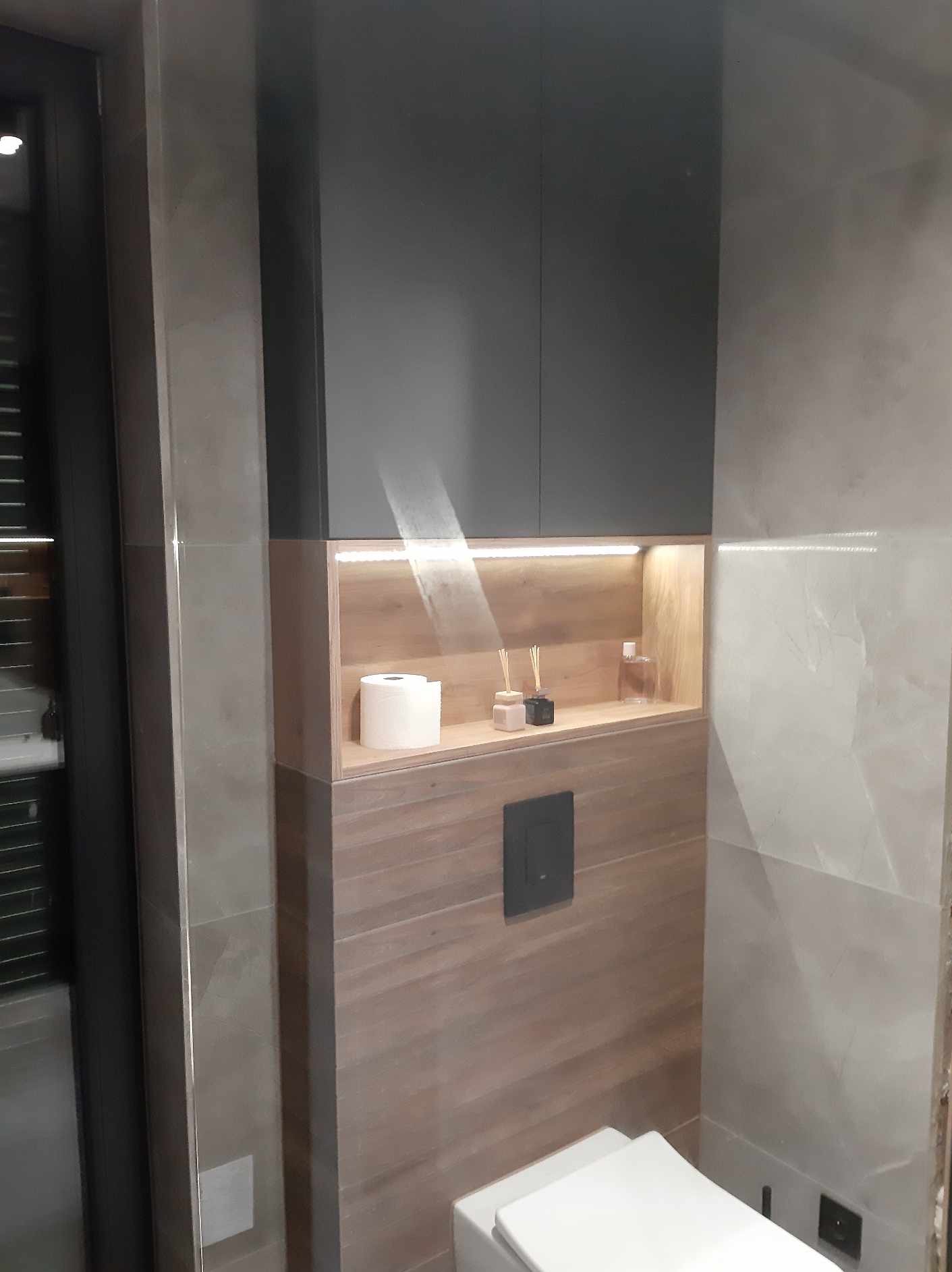 Nowoczesna łazienka w Sulikowie z zabudową WC: drewnopodobne panele ścienne, szara szafka wisząca, podświetlana półka z dekoracjami i rolką papieru toaletowego.