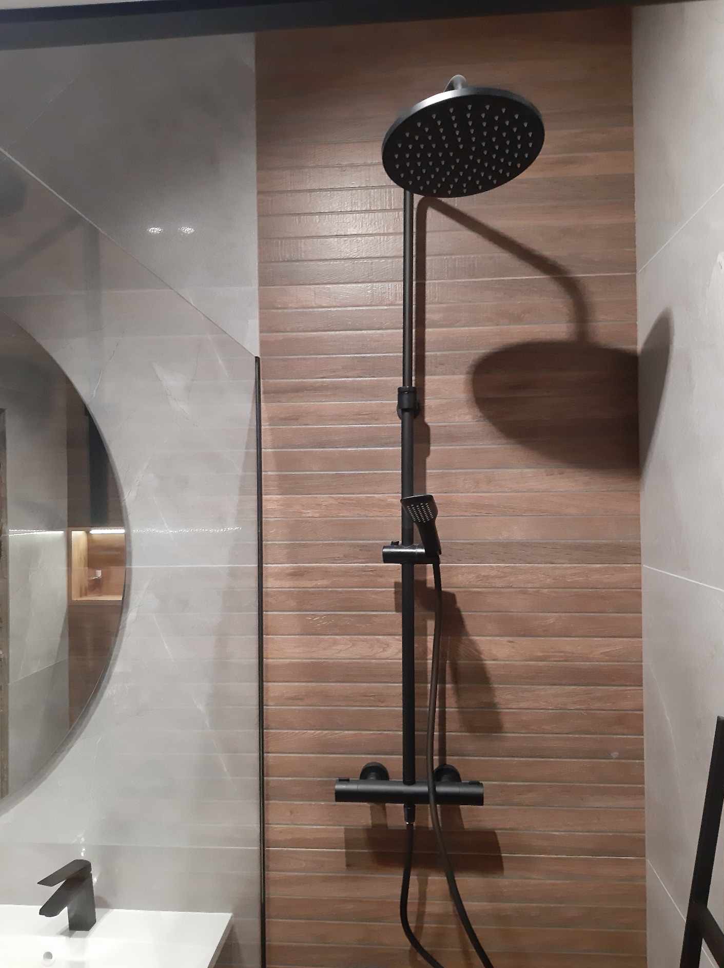 Nowoczesna kabina prysznicowa z czarną armaturą na tle ściany wyłożonej brązowymi płytkami imitującymi drewno, obok umywalka z lustrem.