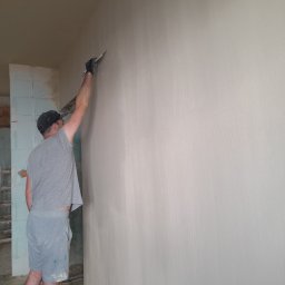 ZAHIDBUD VIKTOR IVASIV - Pracownik w szarym ubraniu wygładza świeżo nałożoną warstwę gładzi na ścianie, używając szpachelki; widoczne nierówności i faktura na powierzchni ściany.