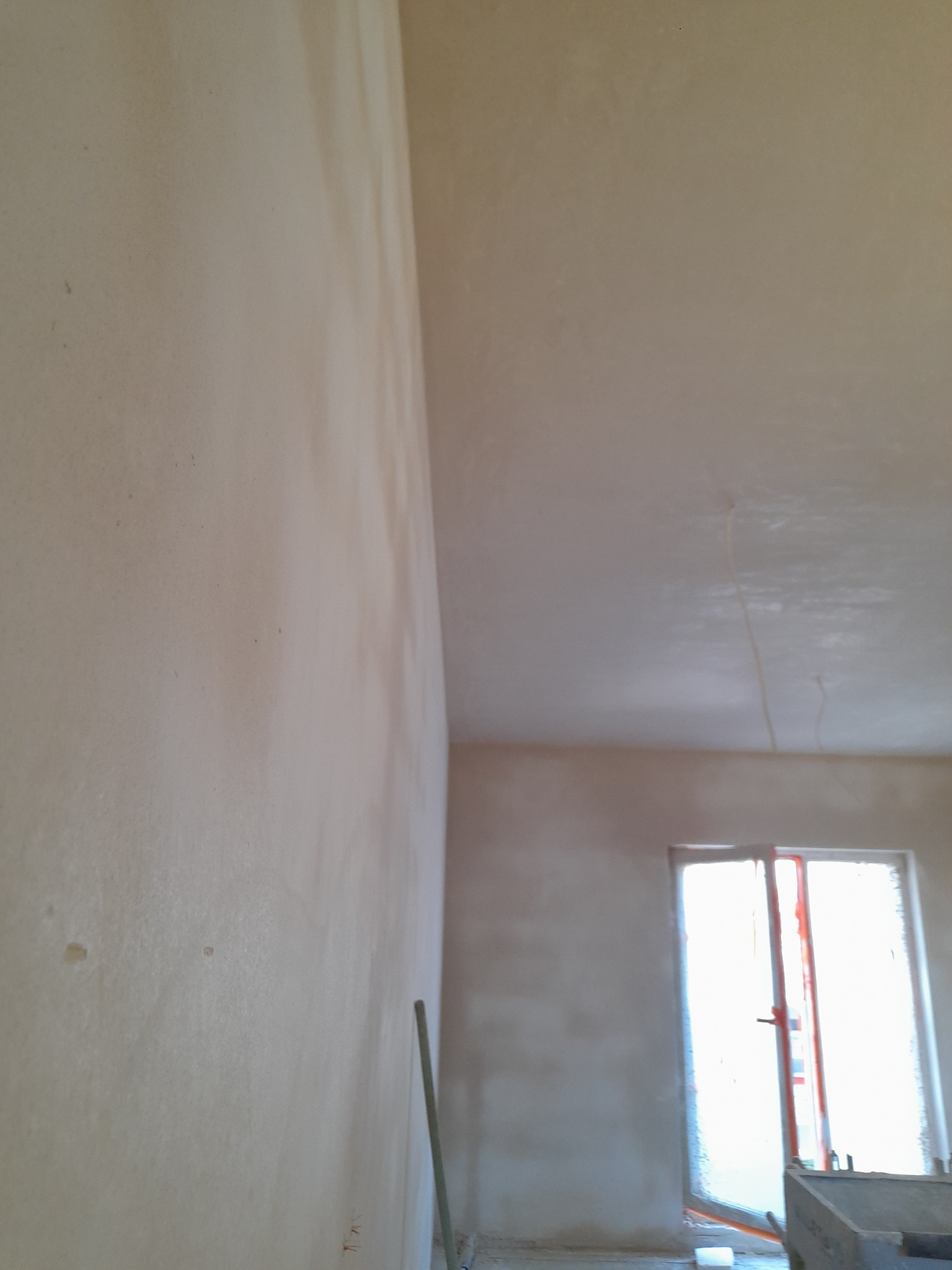 Świeżo nałożony, jasny tynk na ścianach i suficie wewnątrz pomieszczenia w trakcie prac wykończeniowych. Widoczne ślady po narzędziach i delikatne nierówności na powierzchni. W oddali okno...