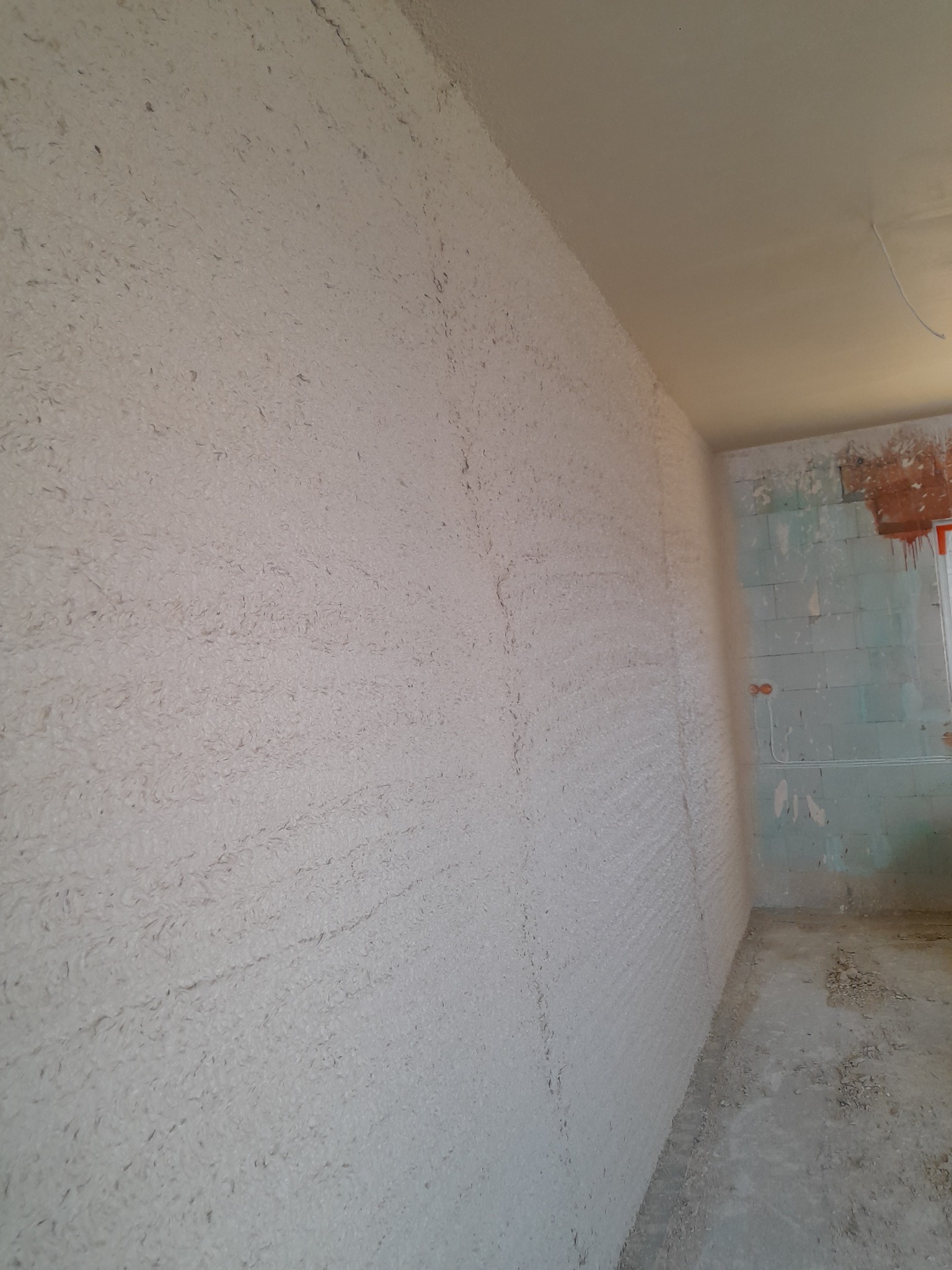 Świeżo nałożony, surowy tynk maszynowy na ścianie w rogu pomieszczenia, widoczne niedoskonałości i nierówności faktury, surowy stan wykończenia wnętrza.