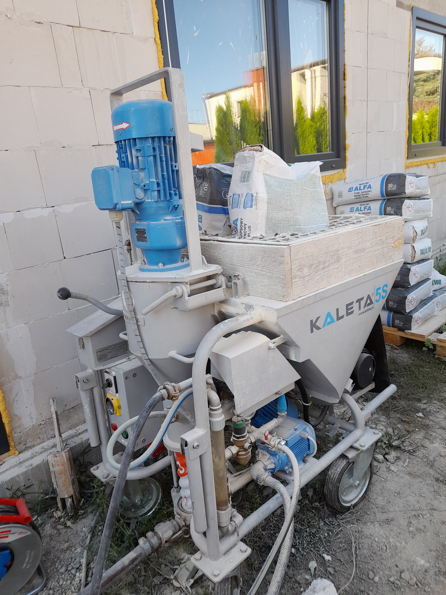 Agregat tynkarski Kaleta 5S z zapasem worków z gipsem Alfa 30kg, stojący na zewnątrz budynku z białego bloczka, widoczne okna z odbiciem zieleni.