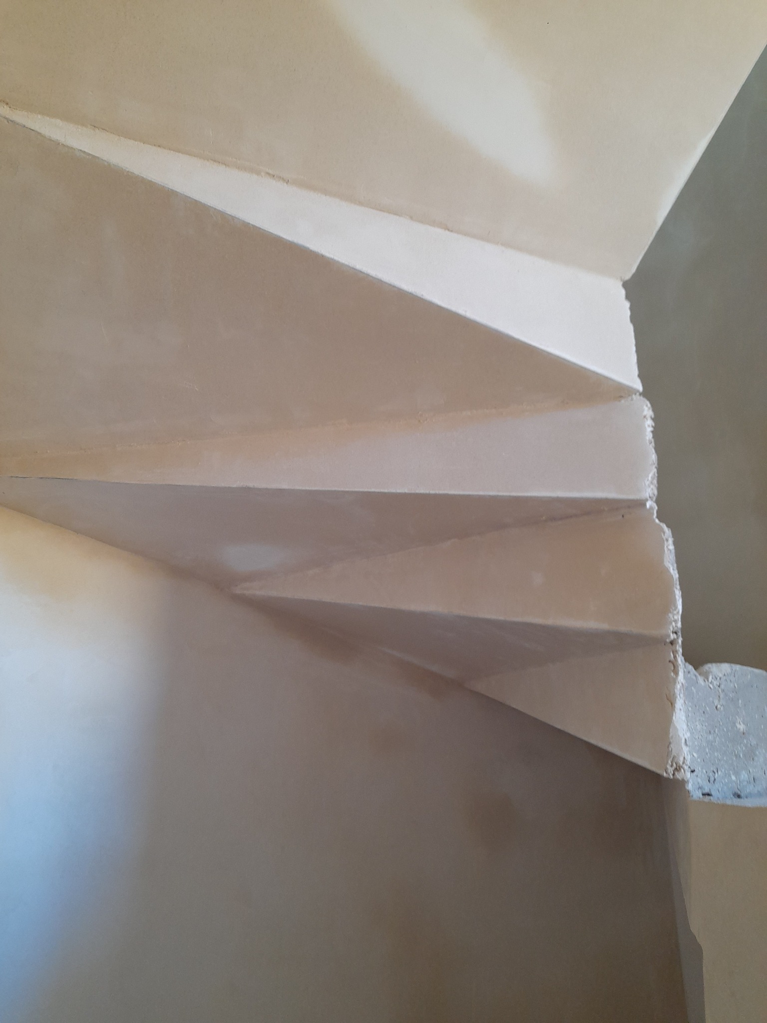 Spiralne schody w trakcie prac wykończeniowych, widoczne świeżo położone tynki w jasnym odcieniu beżu, surowe krawędzie stopni wymagające dalszej obróbki.