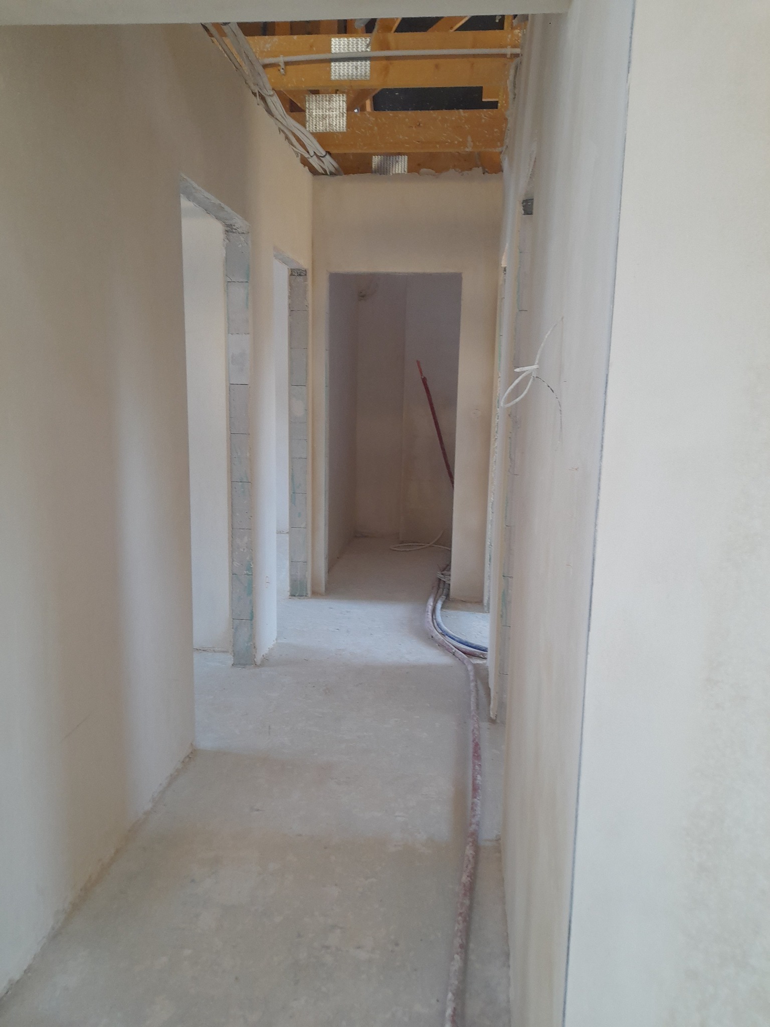 Korytarz w trakcie prac wykończeniowych, widoczne ściany po nałożeniu gładzi, niezabezpieczone kable i przewody elektryczne wystające ze ścian i sufitu, oraz surowy strop z drewnianych belek.