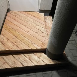 Nazarii Sheketa - Dwustopniowe, drewniane schody wejściowe z desek ułożonych ukośnie, oświetlone z góry, z widoczną kolumną i zwiniętym przedłużaczem w tle.