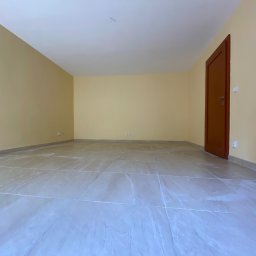 Nazarii Sheketa - Puste, jasne pomieszczenie po remoncie z beżowymi ścianami, nową, jasną, połyskującą podłogą z płytek imitujących kamień, brązowymi drzwiami po prawej stronie i białym grzejnikiem z niebieską...