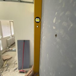 Nazarii Sheketa - Żółta poziomica przyłożona do szarej ściany z widocznymi śladami po szpachlowaniu, w tle fragment pokoju z oknem i grzejnikiem oraz płyta kartonowo-gipsowa oparta o ścianę.