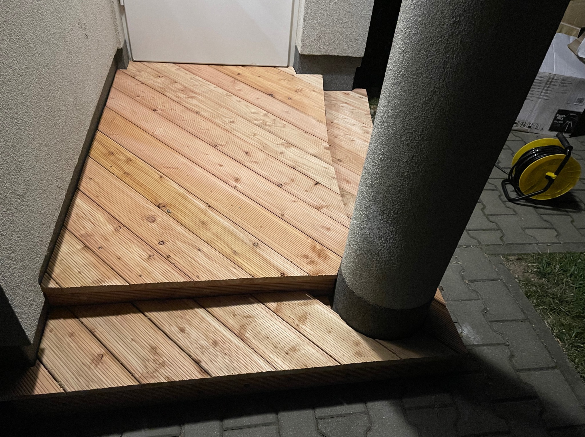 Dwustopniowe, drewniane schody wejściowe z desek ułożonych ukośnie, oświetlone z góry, z widoczną kolumną i zwiniętym przedłużaczem w tle.