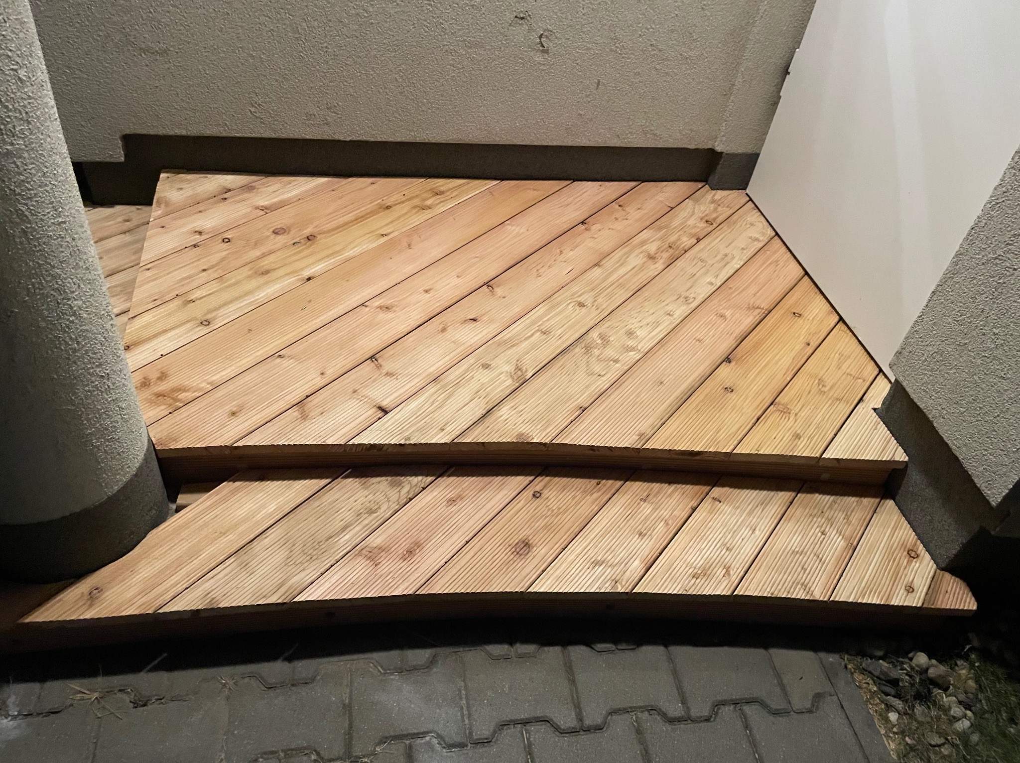 Nowo ułożone, jasne panele drewniane tworzące schody wejściowe, ułożone pod kątem, widoczne szare płyty chodnikowe na dole kadru.