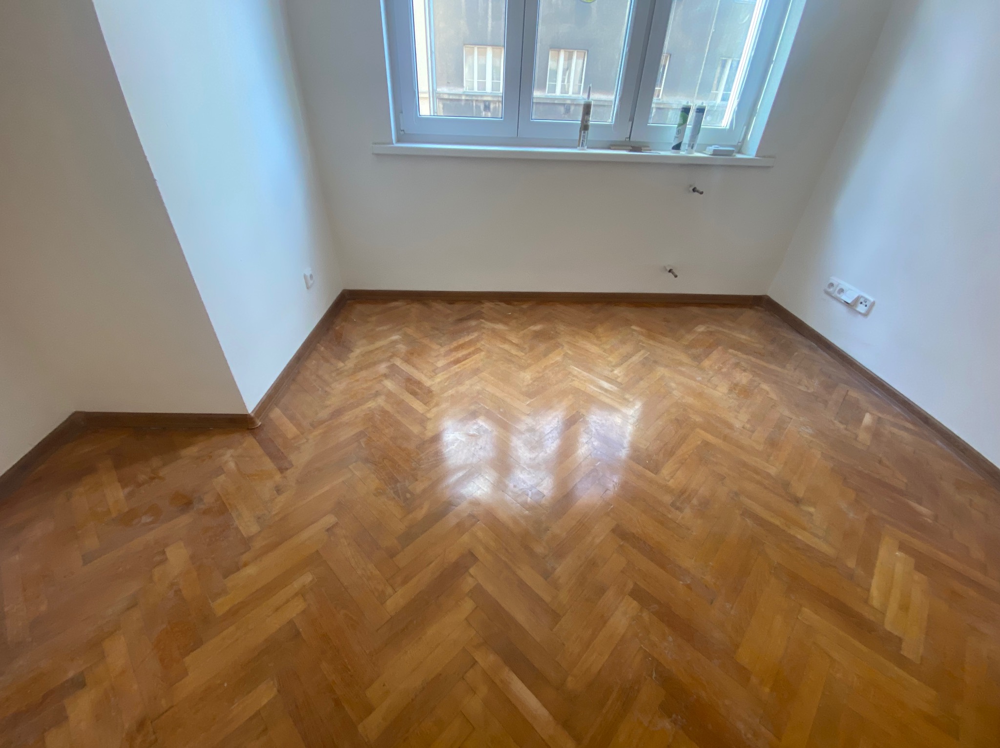 Pusty pokój z parkietem ułożonym w jodełkę, świeżo pomalowane ściany na biało, okno z widokiem na budynek mieszkalny.