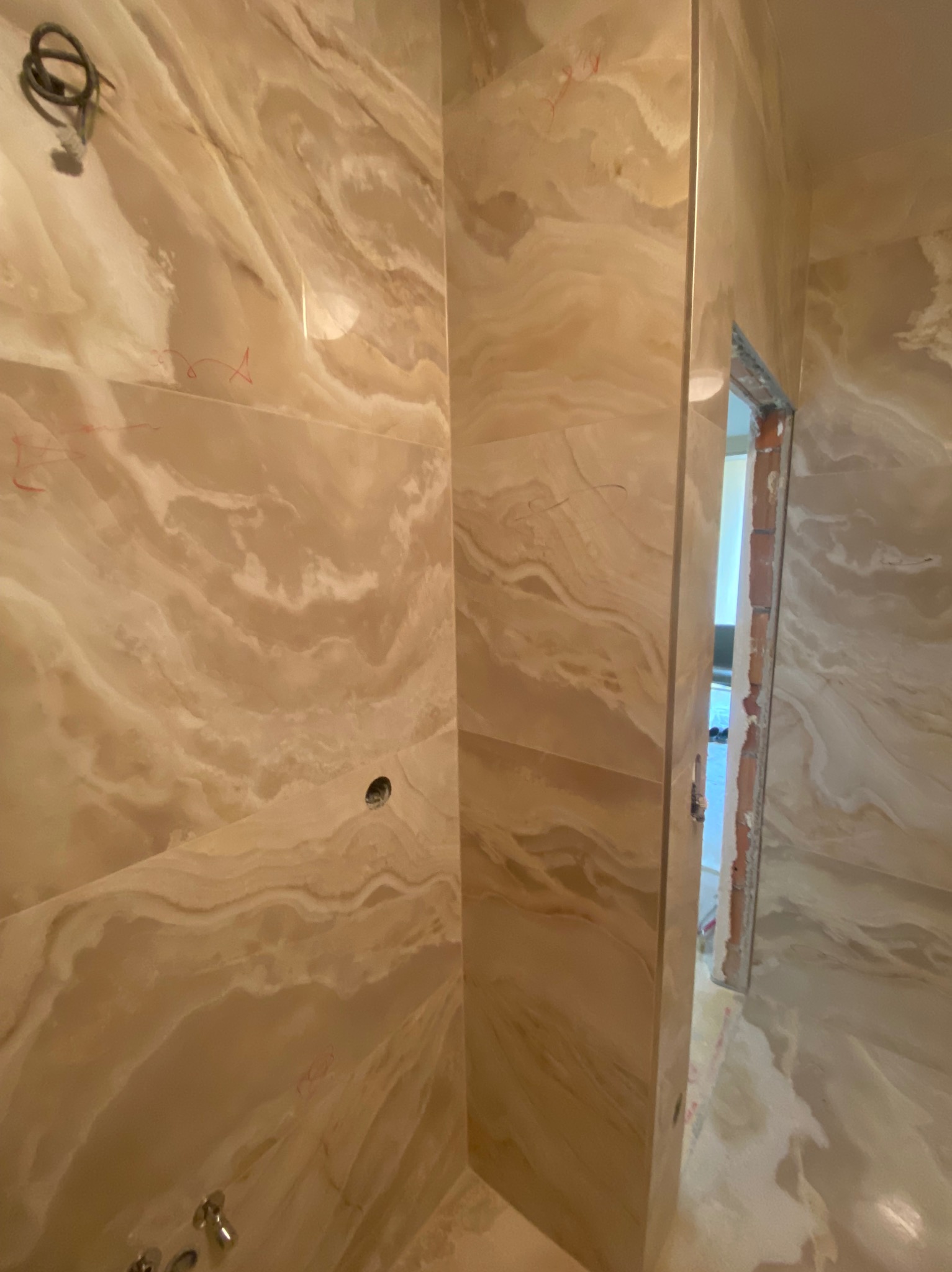 Ściany łazienki wyłożone beżowymi płytami imitującymi marmur, widoczne ślady po ołówku, wystające kable i otwór w ścianie pod instalację, surowy stan wykończenia ościeżnicy.