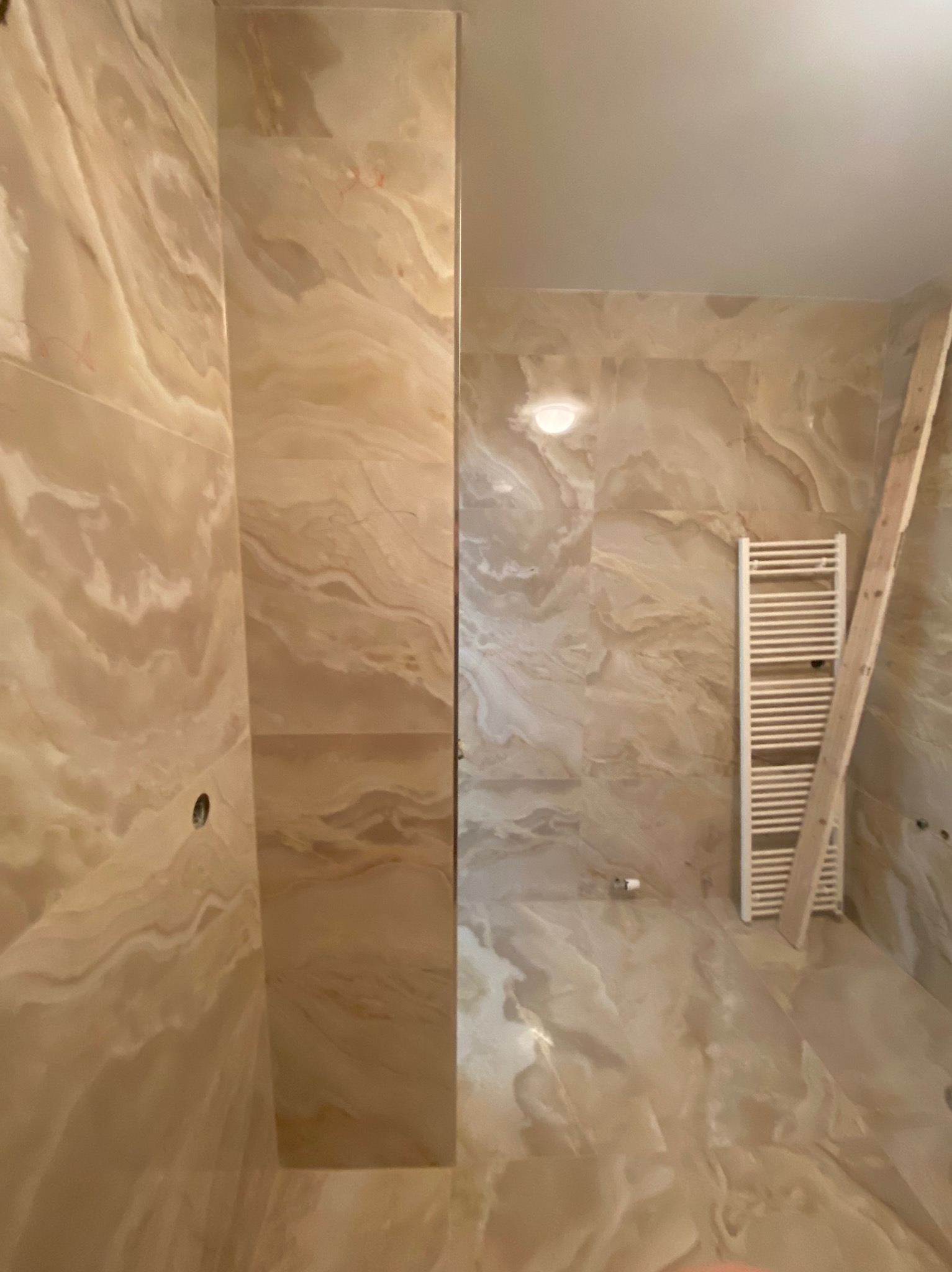 Wykończona łazienka z beżowymi płytkami imitującymi marmur na ścianach i podłodze, widoczny grzejnik drabinkowy i drewniana drabina oparta o ścianę.