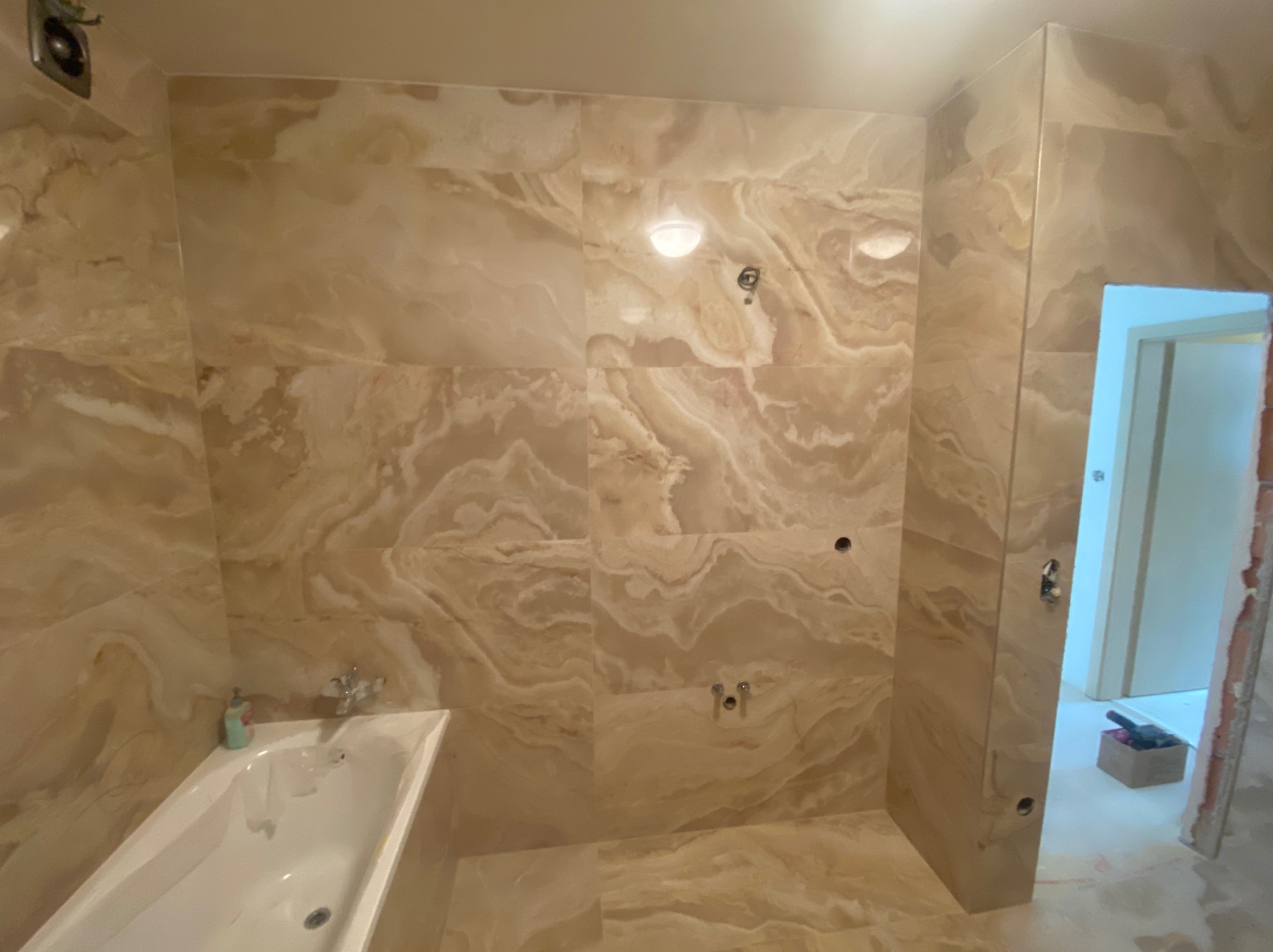 Wykończona łazienka z beżowymi płytkami imitującymi marmur, widoczna wanna, otwory pod armaturę, częściowo otwarte drzwi do sąsiedniego pomieszczenia z narzędziami.