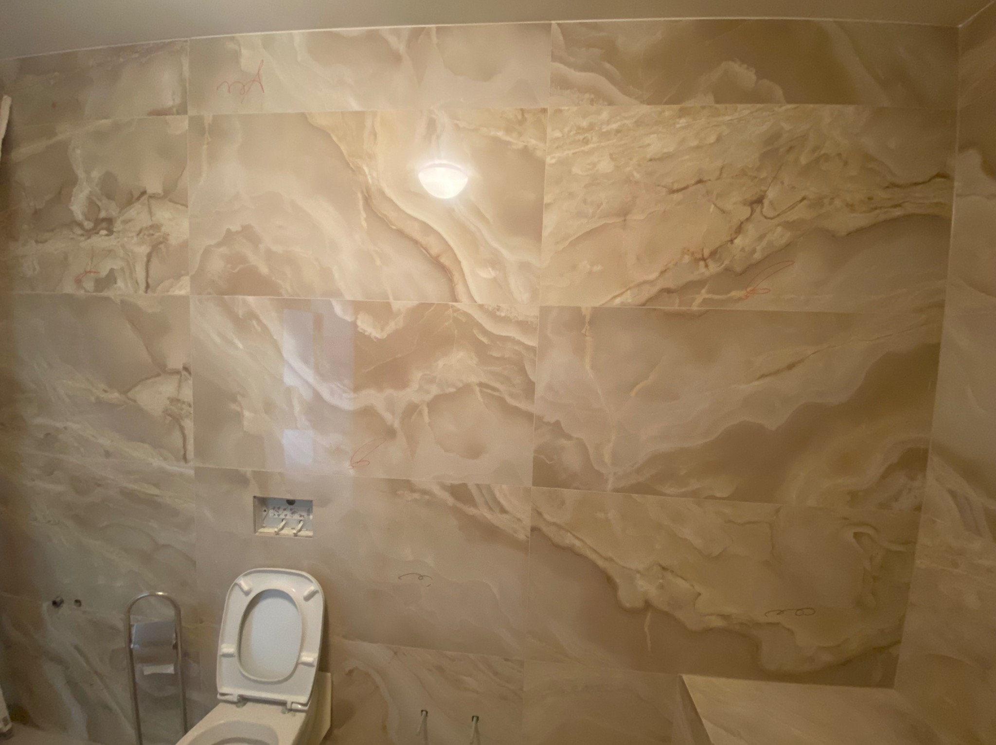 Łazienka w trakcie remontu: widoczne płytki imitujące marmur na ścianach, częściowo zamontowana toaleta, ślady ołówka na płytkach i drabinka.