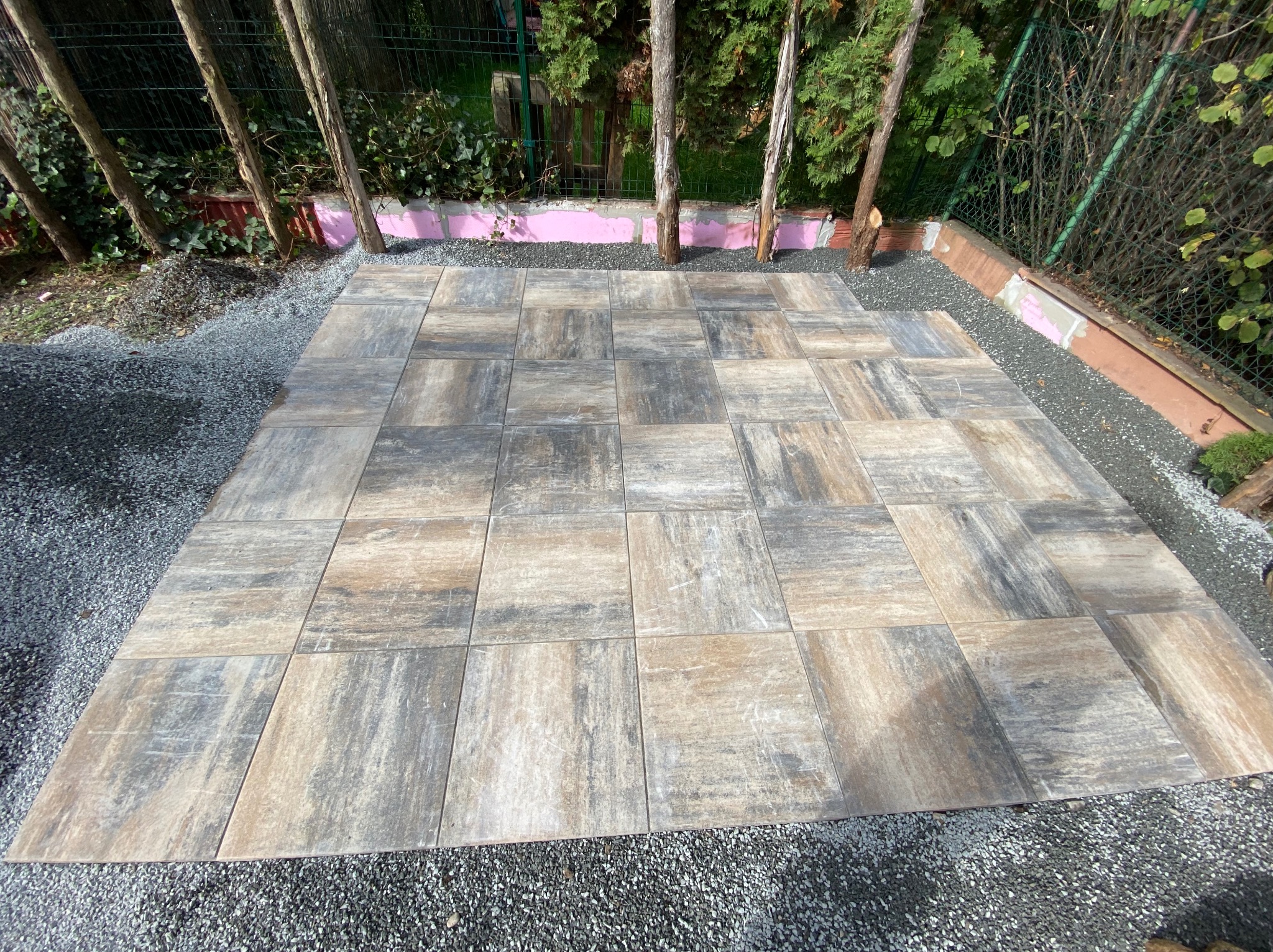 Ułożona nawierzchnia z kwadratowych płyt imitujących drewno na podłożu z drobnego grysu, widoczne drzewa i fragment ogrodzenia w tle.